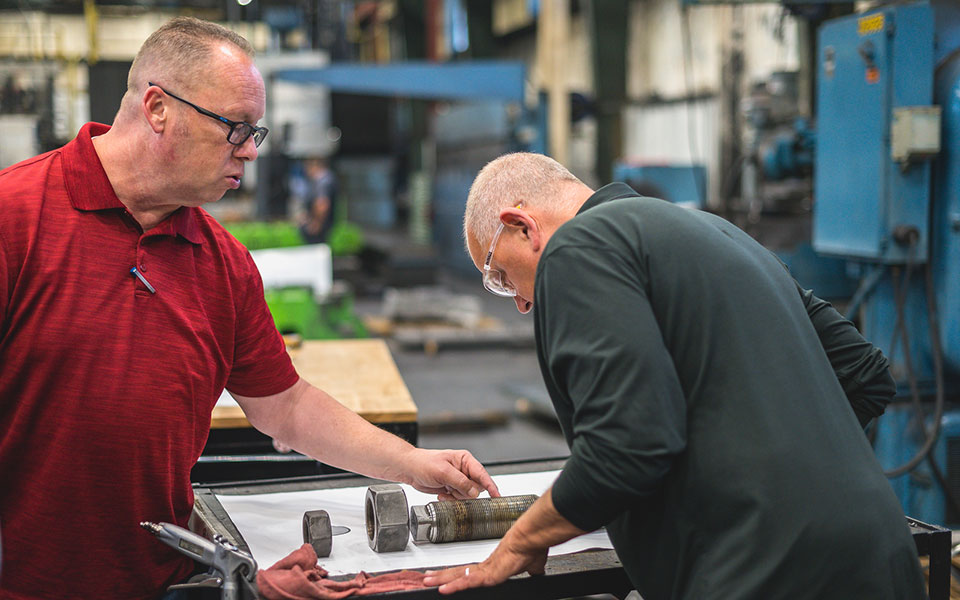 Two employees inspect a round machined piece. One is pointing.