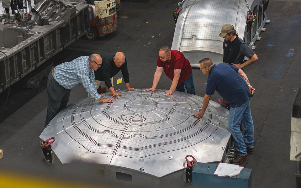 Five employees lean on a very large machined piece with a curved cutout along the top.