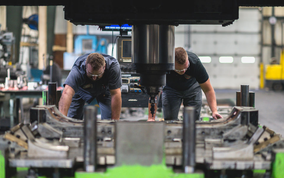 Two employees pull a large, rectangular machined piece away from a large stamping press.