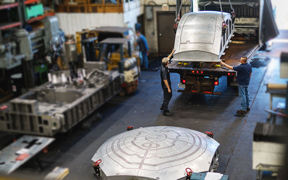 Two employees load a large, metal rounded piece onto the back of a flatbed truck.