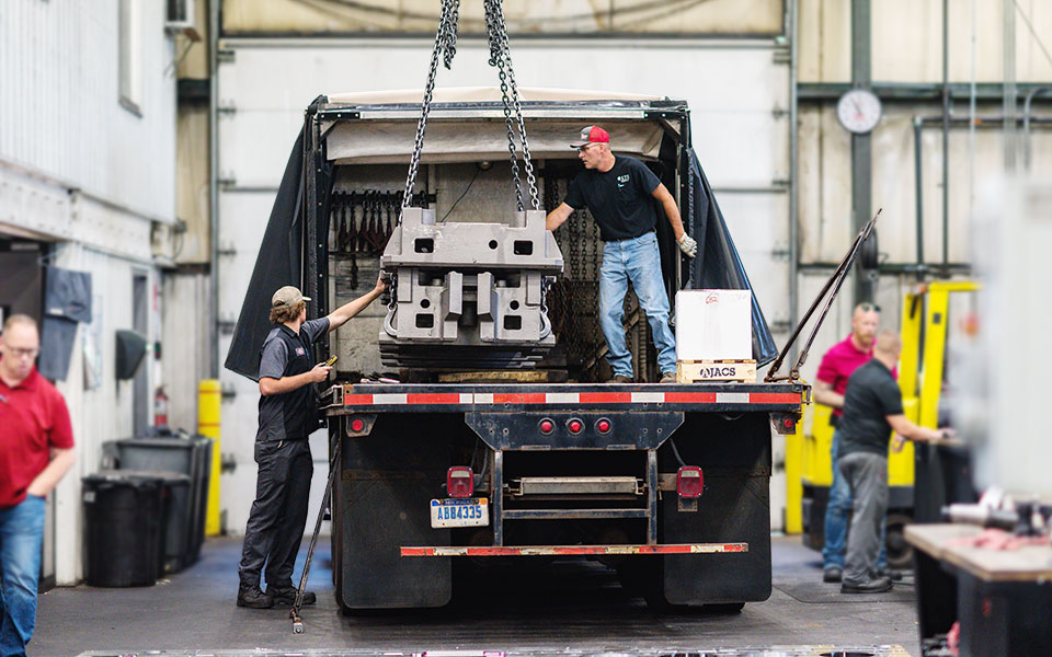 Four employees help load a metal piece onto the back of a large truck.